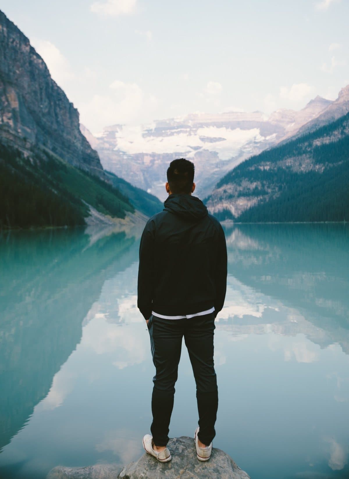 A person standing at the edge of a still mountain lake, looking out at the water